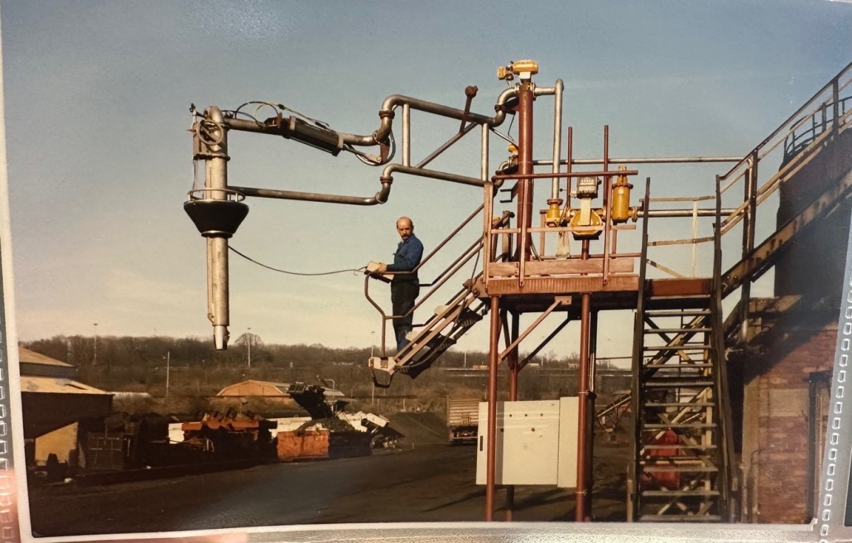 Een man staat op een trap naast een industri&euml;le installatie met metalen buizen en een grote armconstructie, in een fabrieksterrein met stapels grondstoffen op de achtergrond.
