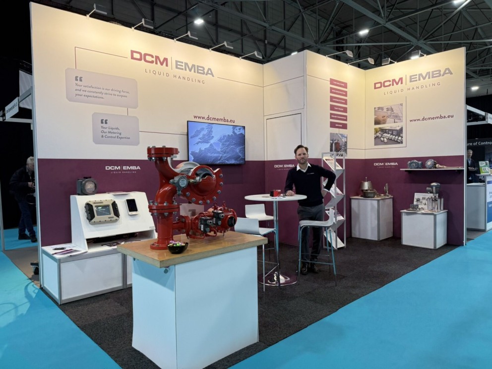 Stand van DCM EMBA op een beurs met liquid handling apparatuur, waaronder een rode pomp, digitale meters en een presentator achter een tafel.