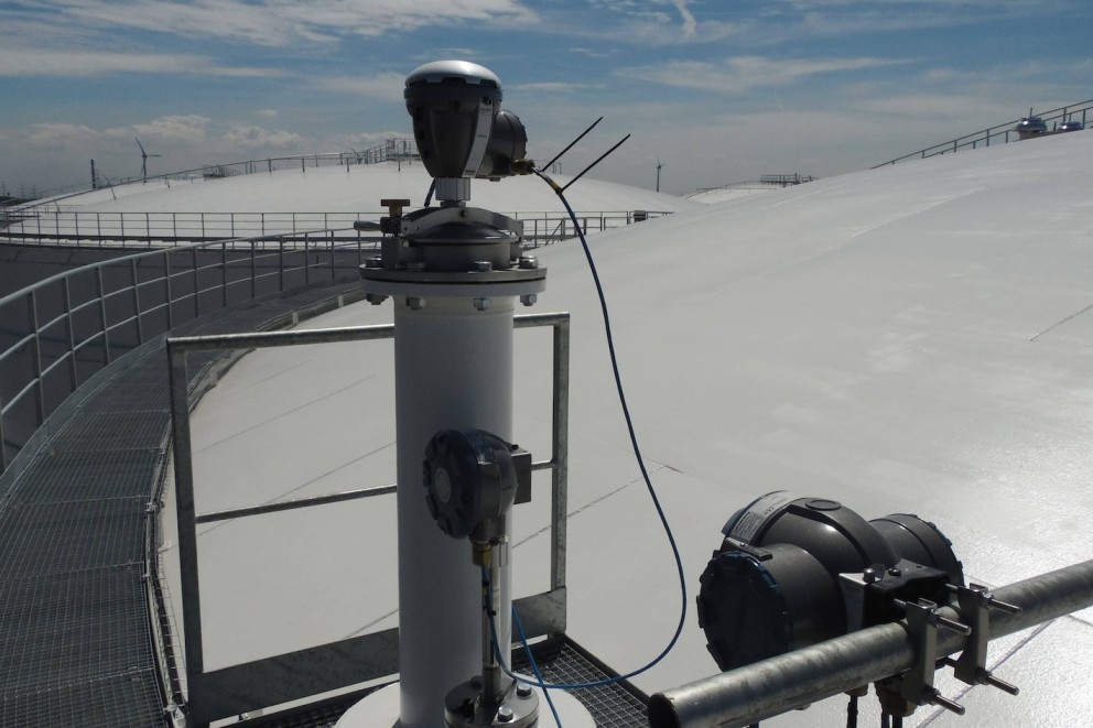 Een industri&euml;le omgeving met een witgekleurde tank en een sensor bevestigd aan een metalen buis, op de achtergrond windturbines en een blauwe lucht met wolken.