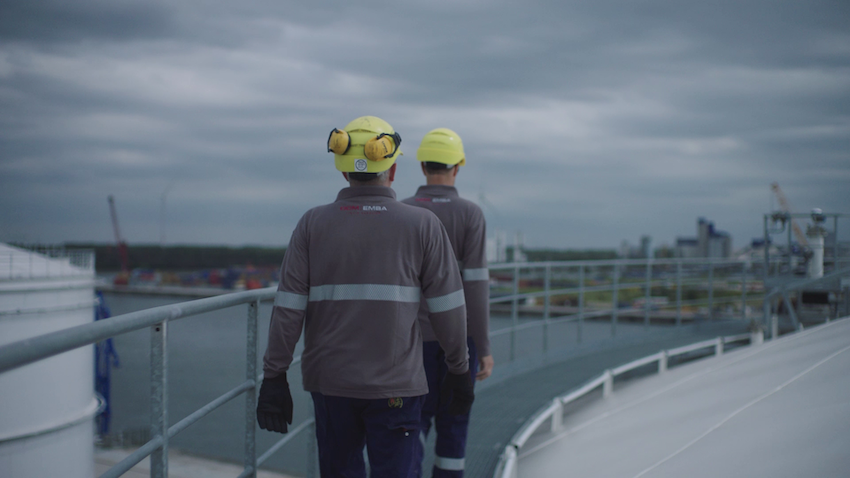 Twee werknemers met veiligheidshelmen lopen over een platform met uitzicht op een haven onder een bewolkte lucht.