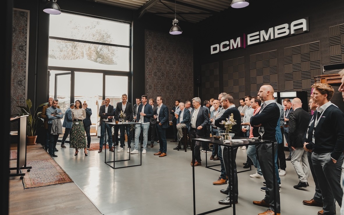Een groep mensen verzameld in een moderne ruimte met grote ramen, terwijl ze naar een spreker kijken; er zijn tafels met drankjes en een paar planten in de ruimte.