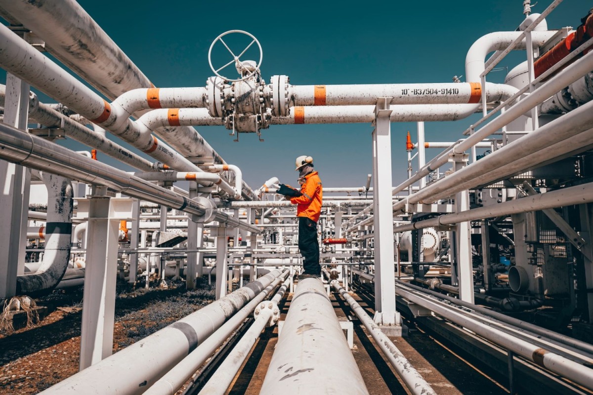 Een werker in een veiligheidspak staat op een pijp tussen verschillende leidingen in een industriële omgeving, terwijl hij aantekeningen maakt.