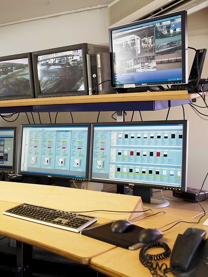 Beeld van een controlekamer met meerdere beeldschermen die camerabeelden en monitoren van een beveiligingssysteem weergeven, met een toetsenbord en muis op een houten bureau.
