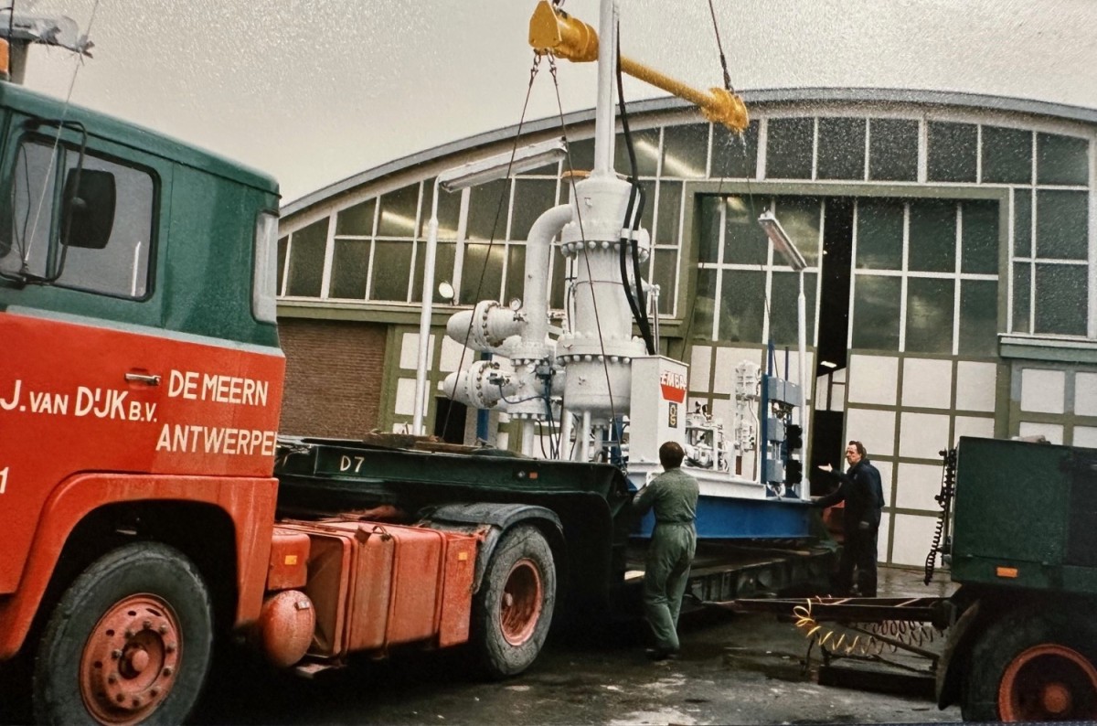 Een vrachtwagen met een wit apparaat wordt gelost bij een loods, terwijl arbeiders aan het werk zijn.