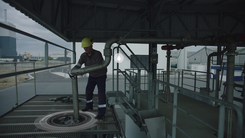 Een werknemer in een gele helm staat op een metalen platform en werkt aan een pijp in een industriële omgeving met een verscheidenheid aan leidingen en tanks op de achtergrond.