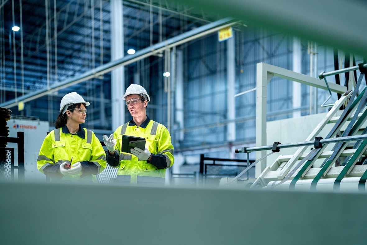 Twee werknemers in veiligheidshesjes en helmen staan in een fabriek en bespreken informatie op een tablet.