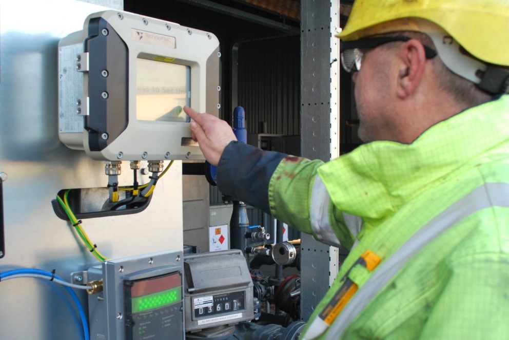 Een werknemer met een veiligheidshelm en een felgekleurde jas bedient een digitaal display van een meetinstrument in een technische omgeving.