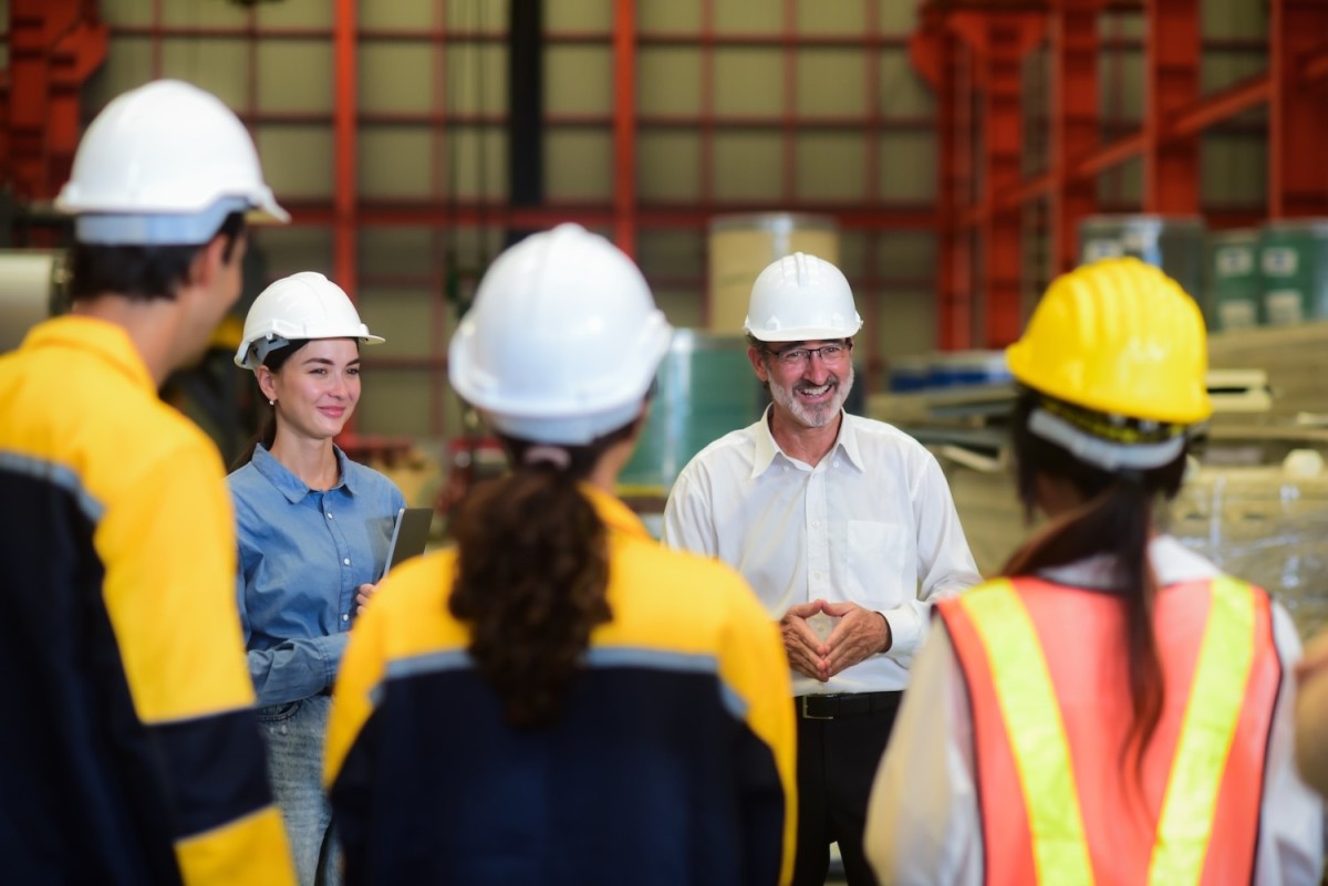 Een groep mensen in veiligheidskleding en helmen staat in een industrieel gebouw, terwijl een man in een wit overhemd en een bril aan het woord is, met een glimlach.