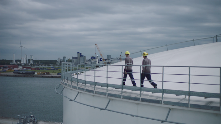 Twee mannen in gele helmen lopen over de rand van een opslagtank met een uitzicht op een haven en een bewolkte lucht.
